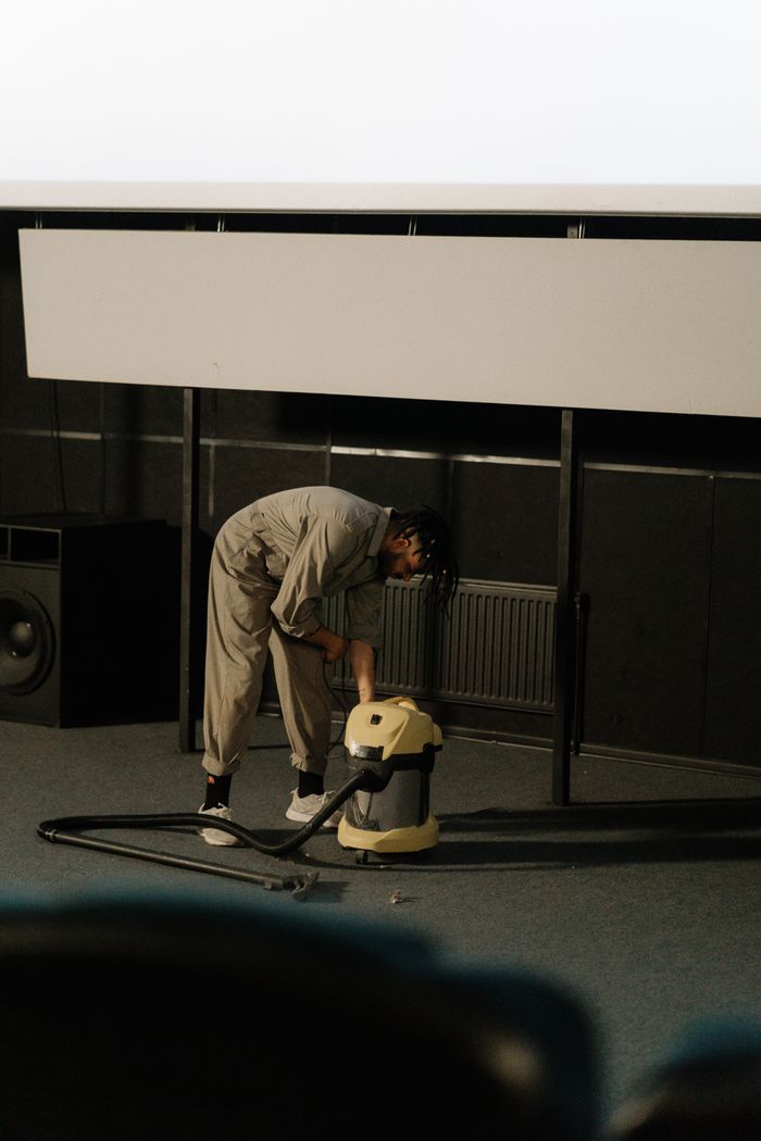 A worker in a jumpsuit using a vacuum cleaner to clean the floor of an auditorium.